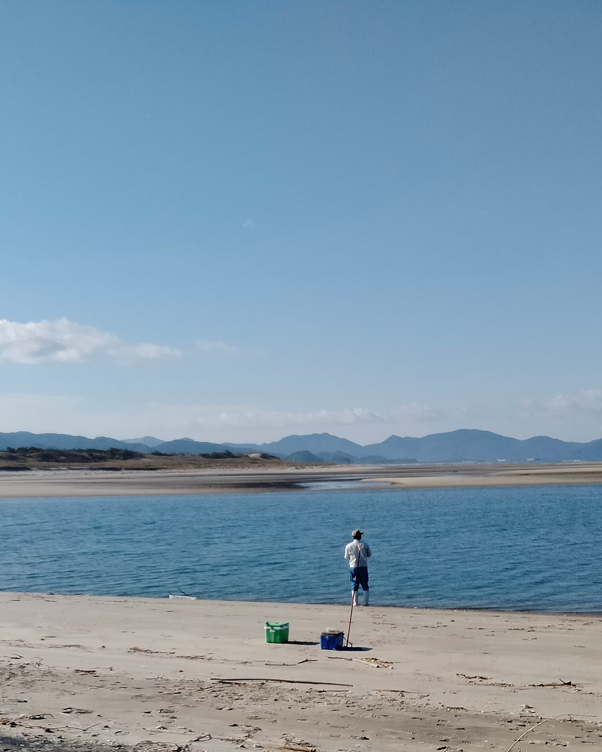 青空の下、穏やかな水辺と遠くの山並みが広がる地元の浜。釣りをする人の後ろ姿